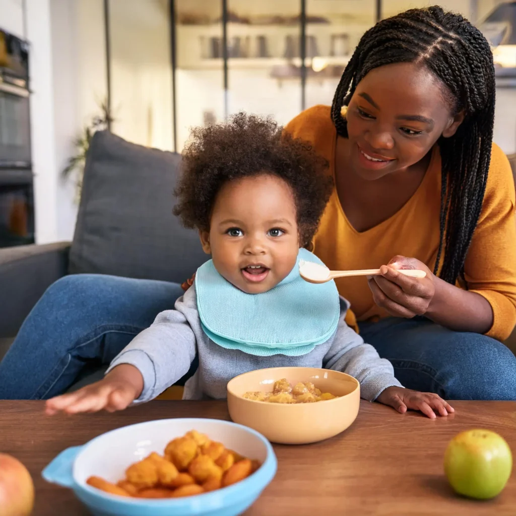 A mother feeding her toddler, who is sitting at a table with a bib, enjoying a meal with bowls of food in front of them.