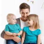 A smiling adult sitting with two young children, one toddler and one older child, both wearing turquoise shirts. The adult holds the toddler while both children exchange smiles.